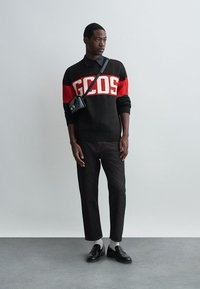 Pullover malha preto e vermelho com o logo "GCDS", calças de ganga pretas, sapatos pretos e meias brancas. O modelo está de pé contra um fundo simples.