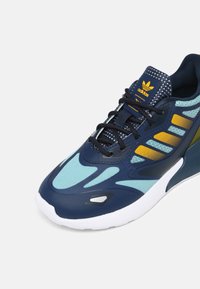 Marinblå atletiska skor med mesh- och syntetöverdel, ljusblå Accenter och gula ränder; vit gummisula med texturerad detalj.