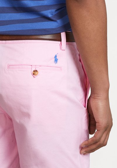 Roza bombažna kratka hlače z zadnjim žepom, ki ima leseno gumb in modro vezeno logotip. Nad temi kratkimi hlačami je oblečena mornarsko modra in modra črtasta majica.