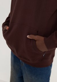 Burgundy sweatshirt with a kangaroo pocket, ribbed cuffs, and a small logo tag on the hem, paired with blue denim jeans.