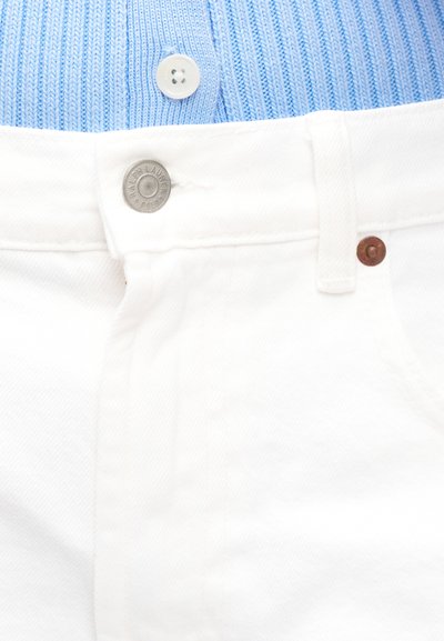 Fehér farmernadrág gomb és cipzár részlet közelről, melyet kék bordázott felső egészít ki, amely deréknál gombolódik össze.