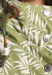 Grönt tyg med vita lövmönster, dekorerat med knappar och en flätad dragsko med färgade pärlor. Sömmarna  är synliga längs kanterna.