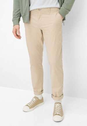 Mann trägt beige Chinos, weiße Socken, beige Turnschuhe, weißes Hemd und grüne Jacke mit Reißverschluss, steht vor weißem Hintergrund.