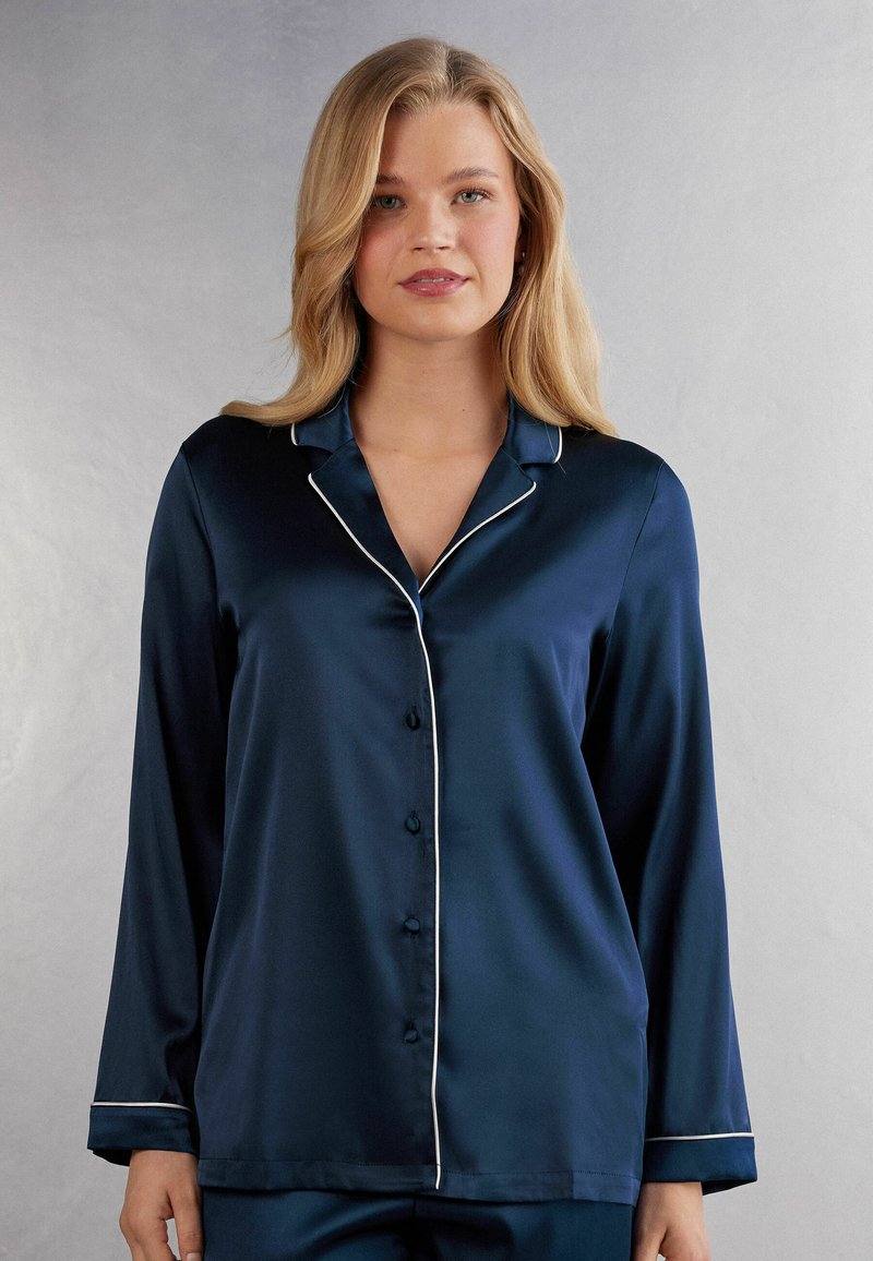 Navy satin pyjamas topp med långa ärmar, en notch-krage, vit pipning och knäppning fram. Slät textur och avslappnad passform.