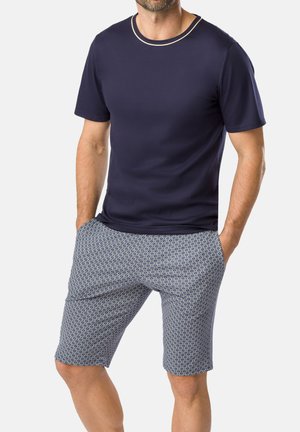 Mann trägt ein dunkelblaues kurzärmeliges Hemd und gemusterte knielange Shorts mit den Händen in den Taschen vor einem einfarbigen Hintergrund.