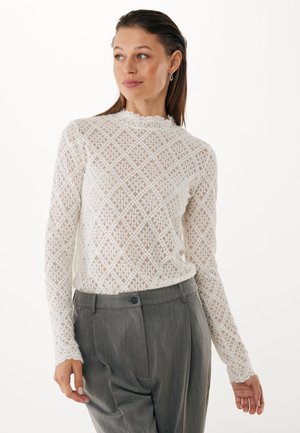 Top de encaje blanco de manga larga con patrones de diamantes, escote de pico con ondas y diseño texturizado. Combinado con pantalones gris ajustados.