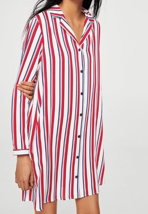 Frau in einem langärmeligen, knöpfbaren Kleid mit vertikalen rot-, weiß- und blauen Streifen auf einem einfarbigen Hintergrund.
