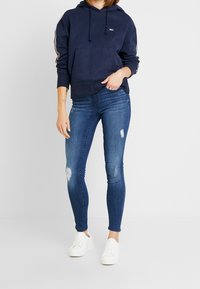 Sudadera navy con bolsillo canguro, cordones blancos y rayas laterales. Combinada con jeans skinny azules desgastados y zapatillas blancas.