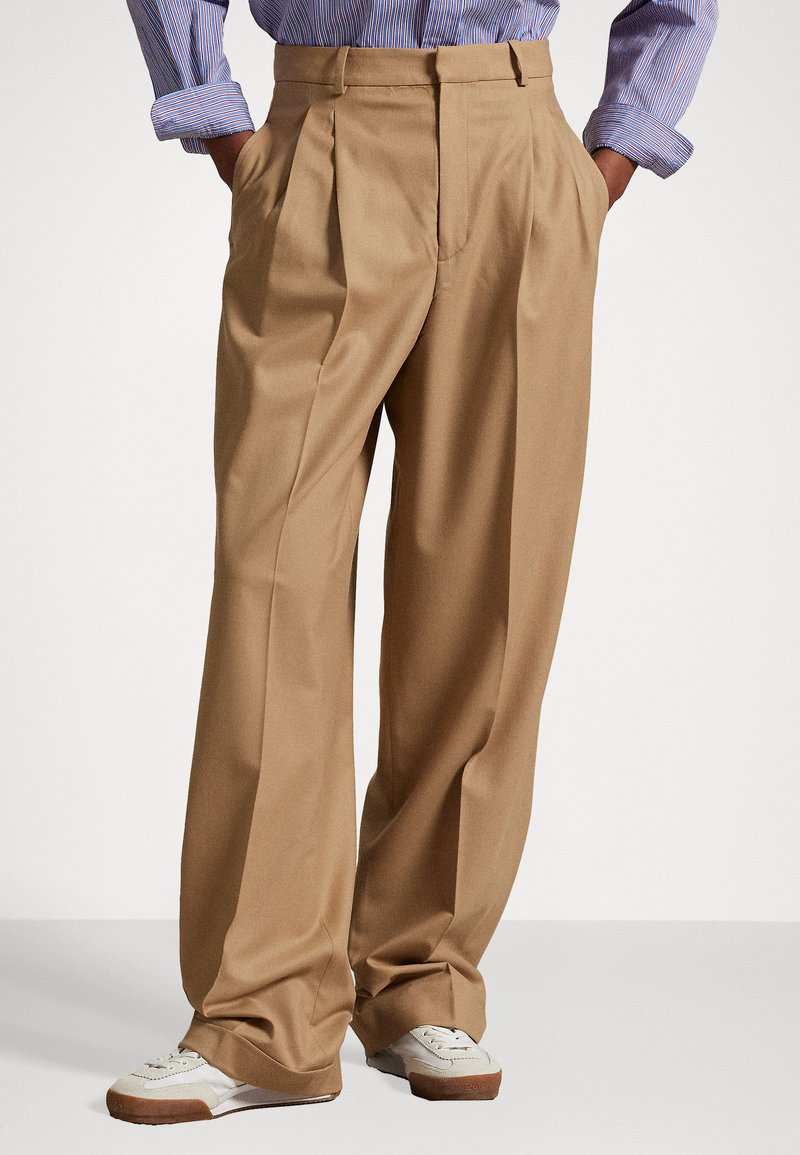 Beige vida byxor med plisseringar, tillverkade av mjukt tyg, med hög midja och skräddarsydd passform, kombinerat med vita sneakers.