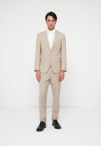 Beige szabott öltöny egy egysoros kabáttal és hozzá illő nadrággal, fehér inggel és fekete bőr cipővel párosítva. Sima textúra.