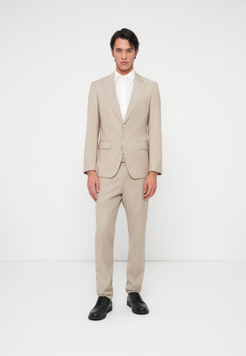 Beige szabott öltöny egy egysoros kabáttal és hozzá illő nadrággal, fehér inggel és fekete bőr cipővel párosítva. Sima textúra.