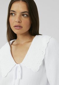 Blouse blanche avec un grand col brodé, fermeture à nouer à l'avant, tissu texturé et doux plissé. Accent sur le détail floral du col.
