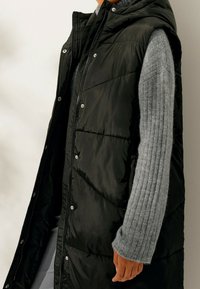 Černá nafouknutá vesta s kapucí, se zapínáním na zip, patentními knoflíky a jemně prošívanými částmi. Nosená přes světle šedý žebrovaný svetr.