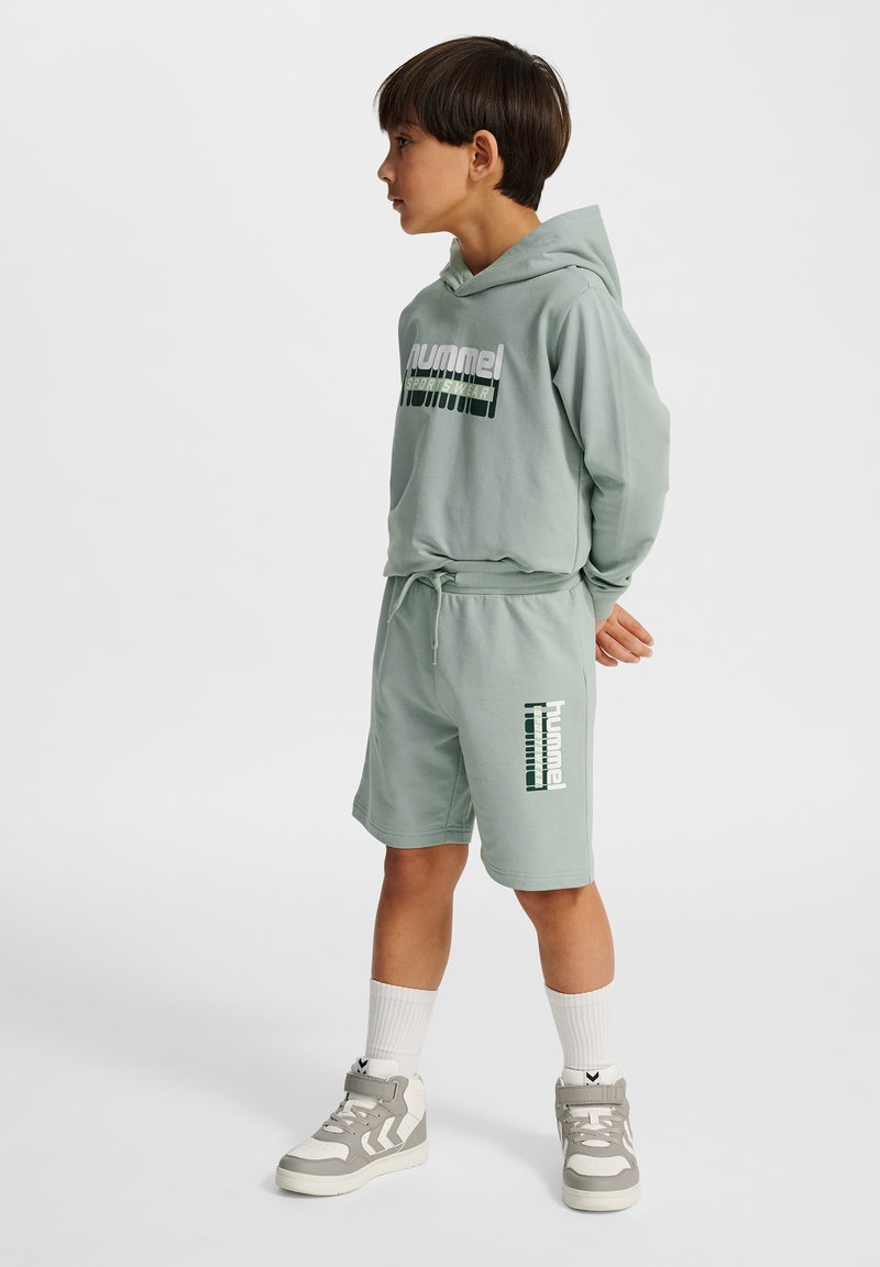 Ljusgrönt set med hoodie och shorts i mjukt material, med logotryck och dragsko i midjan, matchat med grå höga sneakers.