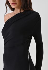 Asymmetrisches schwarzes Kleid aus glattem Stoff, mit Drapierungen und einem dekorativen Reißverschluss an der Seite, der vom Saum verläuft.