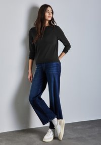 Pull noir à manches longues, jean bleu à jambes larges, baskets blanches avec des accents crème. Le mannequin se tient contre un mur gris clair.