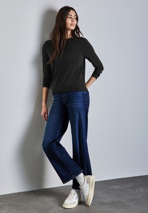 Pull noir à manches longues, jean bleu à jambes larges, baskets blanches avec des accents crème. Le mannequin se tient contre un mur gris clair.