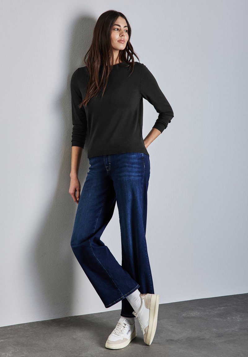 Pull noir à manches longues, jean bleu à jambes larges, baskets blanches avec des accents crème. Le mannequin se tient contre un mur gris clair.