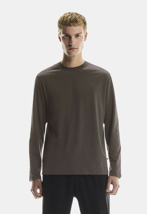 Jonge man met kort blond haar draagt een eenvoudige lange mouwen bruine shirt en zwarte broek, staand tegen een witte achtergrond.