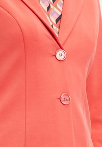 Korallenfarbener Blazer mit glatter Textur, ausgestattet mit zwei runden Knöpfen. Ein gemustertes Hemd ist teilweise unter dem Blazer sichtbar.