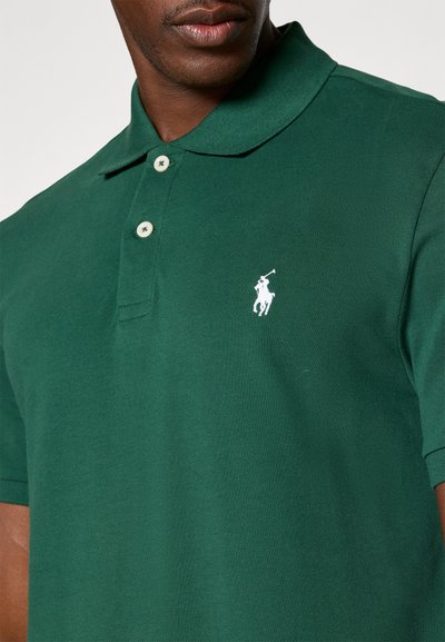 Zaļš polo krekls, kas izgatavots no strukturēta auduma, ar apkakli, trim pogām un baltu izšūtu logotipu kreisajā krūtī.