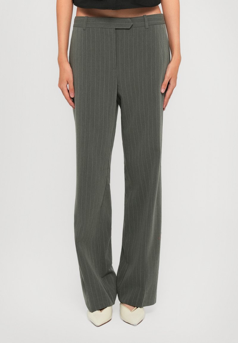 Woman wearing high-waisted, grey pinstripe wide-leg pants with front tab detail and white pointed-toe shoes, standing against a plain background.