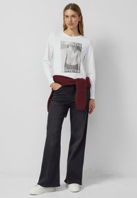 Biała koszula z długim rękawem z nadrukiem graficznym, zestawiona z czarnymi szerokimi spodniami dżinsowymi i bordowym swetrem związanym w talii, białe sneakersy.