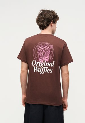 Άνδρας με καφέ μπλουζάκι με ροζ εικονογραφήσεις βάφλας και λευκή επιγραφή "Original Waffles" στην πλάτη, γυρισμένος προς τα πίσω σε απλό φόντο.