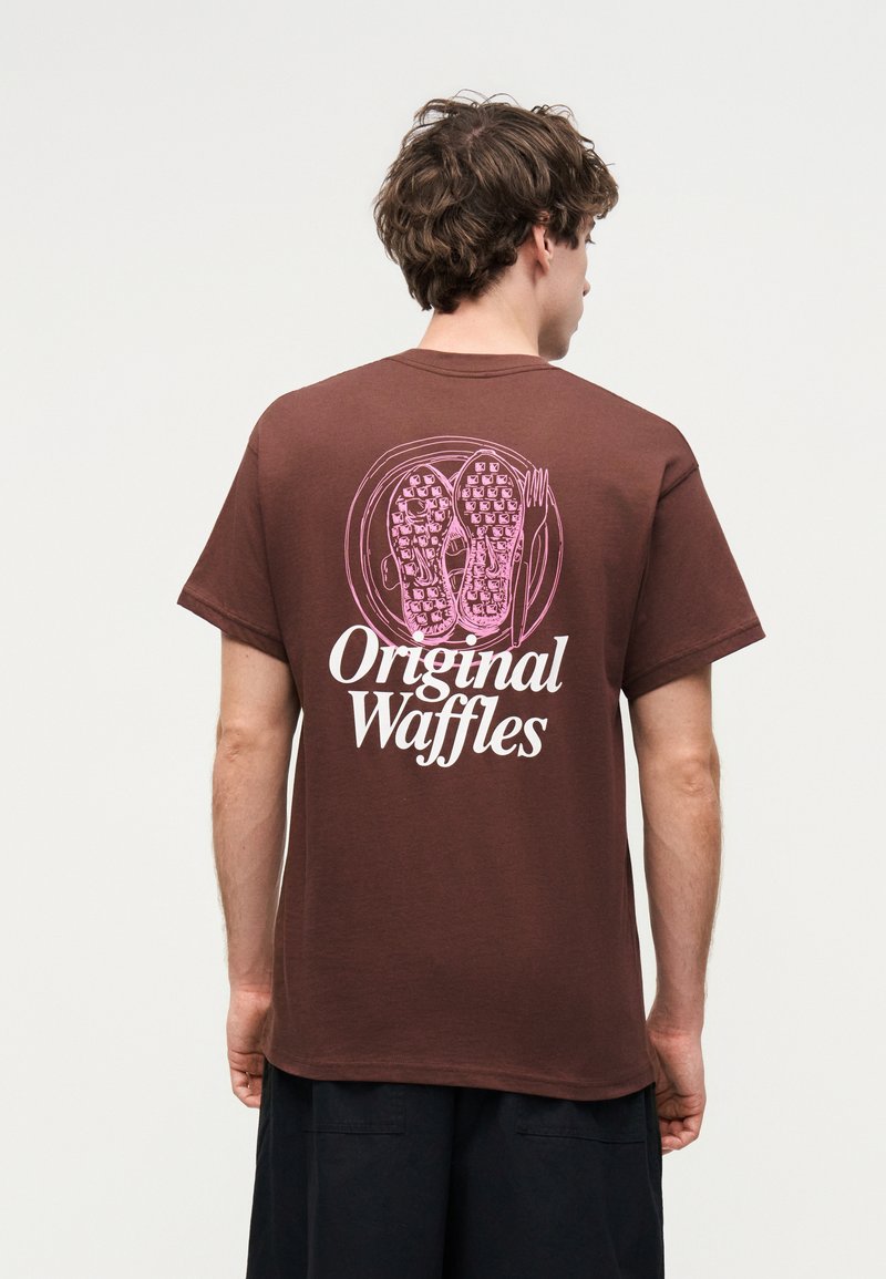 Άνδρας με καφέ μπλουζάκι με ροζ εικονογραφήσεις βάφλας και λευκή επιγραφή "Original Waffles" στην πλάτη, γυρισμένος προς τα πίσω σε απλό φόντο.