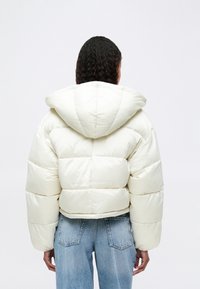 Κοντό μπουφάν σε εκρού, με κουκούλα, καπιτονέ σχέδιο και φαρδιά μανίκια. Συνδυάζεται με ανοιχτό μπλε τζιν παντελόνι.