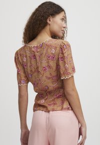 Blusa floreale marrone con maniche a sbuffo corte, bordi scalloppati e motivi intricati in rosa e blu, abbinata a pantaloni rosa chiaro.