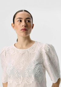 Blouse blanche brodée avec des manches courtes bouffantes, motifs floraux délicats, texture légère et encolure ronde, présentant des accents de couture détaillés.