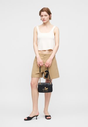 Γυναίκα που φοράει λευκό κοντό τοπ, κοντό σορτς ταμπά μέχρι τα γόνατα, μαύρα σανδάλια με τακούνι, κρατώντας μια μαύρη τσάντα με σχέδιο καπιτονέ και χρυσές αλυσίδες.