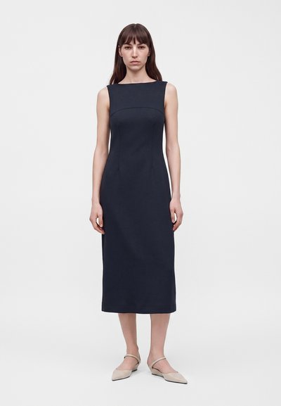 Femme aux cheveux bruns raides, portant une robe midi sans manches bleu marine foncé et des ballerines pointues beige, debout devant un fond blanc uni.