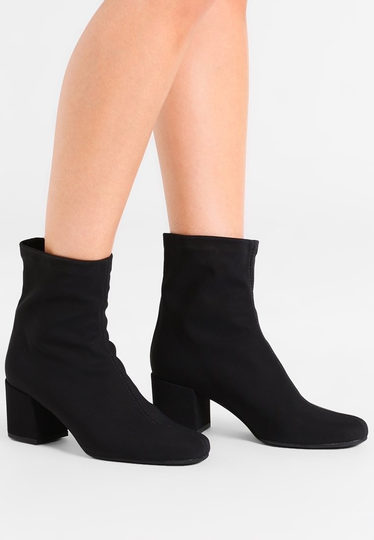 Botines negros ajustados con tacones de bloque, llevados sobre piernas al desnudo, mostrados contra un fondo claro y liso.