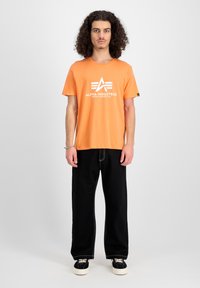 Oranges Baumwoll-T-Shirt mit Rundhalsausschnitt und weißem Bedruck. Kombiniert mit schwarzen, weit geschnittenen Hosen und schwarzen Sneakers mit beigen Schnürsenkeln.