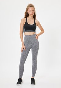 Czarny biustonosz sportowy z szarymi akcentami, zestawiony z wysokimi, teksturowanymi szarymi legginsami. Model nosi czarne buty sportowe.