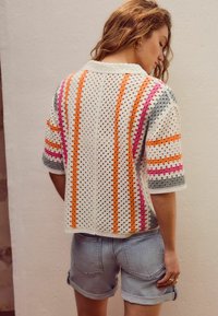 Haut au crochet blanc avec des motifs rayés colorés en orange, rose et gris. Présente des manches courtes et une coupe décontractée. Associé à un short en jean.