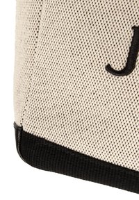 Beżowa torba na ramię z płótna z czarną dolną lamówką i wyhaftowaną literą 'J'. Materiał o fakturze, solidna konstrukcja.