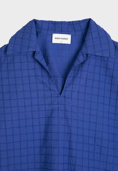 Chemise bleue avec motif quadrillé texturé, col ouvert et étiquette de marque visible "BOBO CHOSES" à l'intérieur du col.