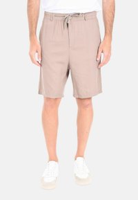 Pantaloni corti beige con vita regolabile, tasche laterali, taglio sartoriale e texture del tessuto liscia. Abbinati a sneakers bianche.