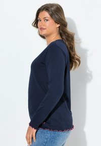 Modrá dlouhá halenka s kulatým výstřihem a růžovým vlnitým lemem. Tkanina je hladká a je kombinována s džíny.