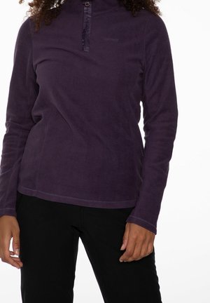 Person iført en langærmet mørkelilla fleecepullover med en kort lynlås og sorte bukser, stående mod en hvid baggrund.