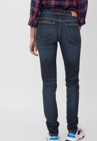 Vaqueros ajustados de color azul oscuro con una textura suave, bolsillos traseros y costuras en contraste. La cintura presenta una etiqueta de cuero marrón.