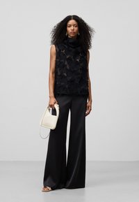 Top nero senza maniche con dettagli a frange, abbinato a pantaloni neri a gamba larga. Tiene una piccola borsa bianca con forma unica.