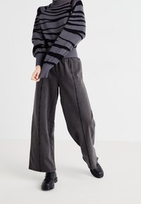 Pull en maille à col roulé rayé gris avec accents noirs, assorti à un pantalon gris foncé à jambes larges. Les deux présentent des matériaux doux et des finitions texturées.