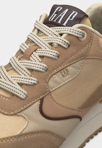 Μπεζ και καφέ αθλητικό παπούτσι με λευκά και καφέ ραμμένα κορδόνια, υφασμάτινες πάνελ με υφή και τη σήμανση «GAP» στη γλώσσα και στην πλευρά.