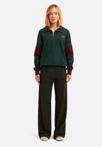 Felpa verde con zip e dettagli bordeaux sulle maniche, abbinata a pantaloni neri a vita alta e a gamba larga. Tessuto liscio con polsini a coste.