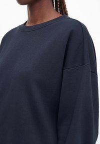 Gros plan d'une personne portant un sweat-shirt bleu marine foncé oversize à col rond, avec des cheveux tressés visibles au niveau du cou.