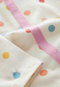 Kokvilnas audums ar krēmkrāsas fonu un krāsainiem punktiņiem dzeltenā, oranžā, rozā, zilā un violetā krāsā. Rozā lentes rotājums.